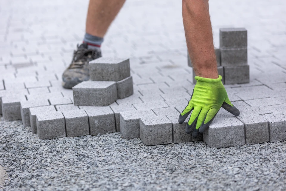 Ouvrier en gants jaunes installant des pavés en béton sur lit de sable, agenouillé sur chantier extérieur