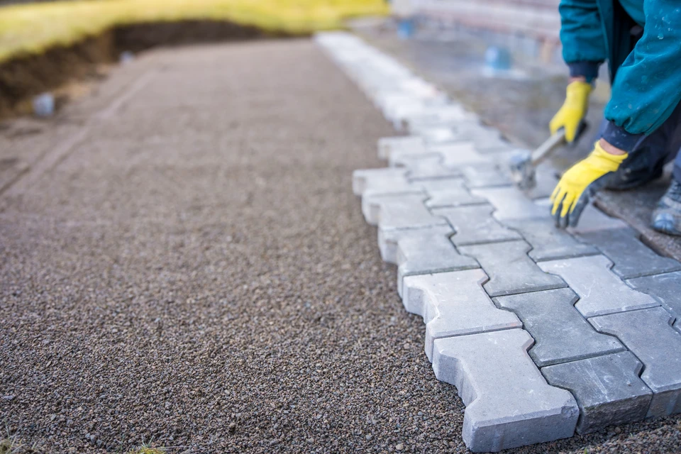 Ouvrier en tenue de travail installant un pavage extérieur, agenouillé sur surface bétonnée avec marteau et gants de protection